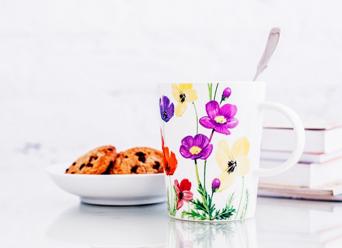 Milchbecher mit Blumenmuster, Kekse und einige Bücher auf Glastisch