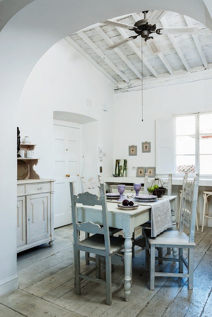 View through arched open doorway into high-ceilinged, grey and white dining room with ceiling fan above set table and antique chairs