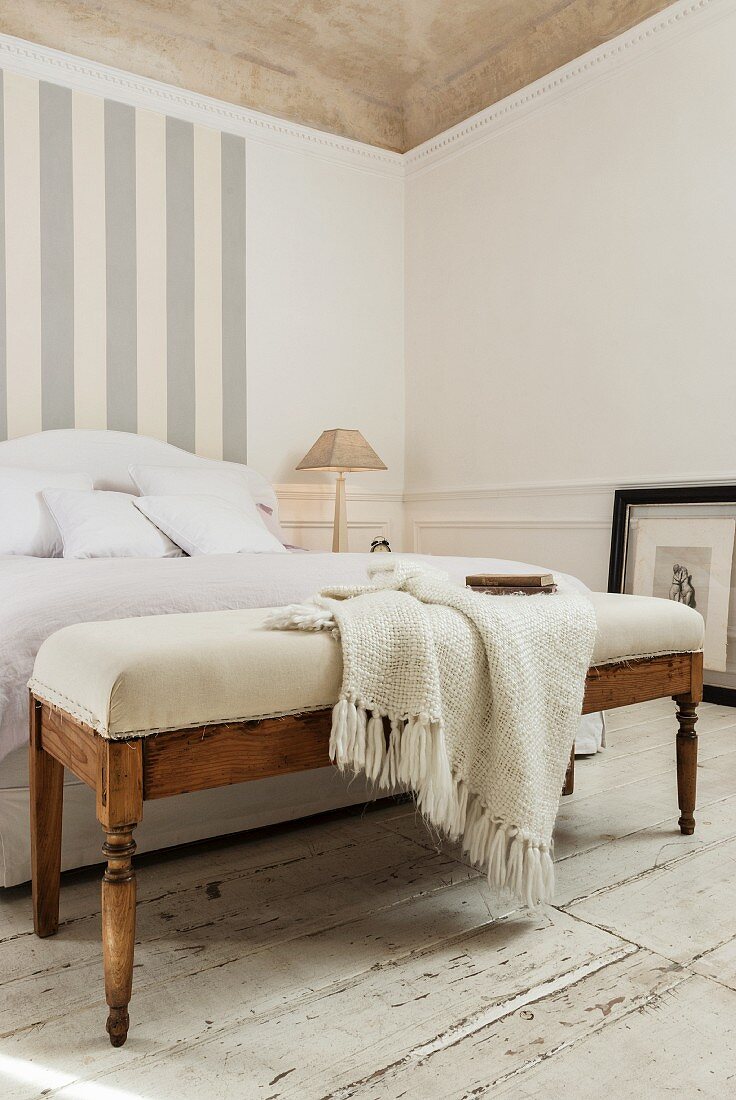 Knitted blanket on upholstered bench at foot of French bed and grey and white pattern of stripes on wall