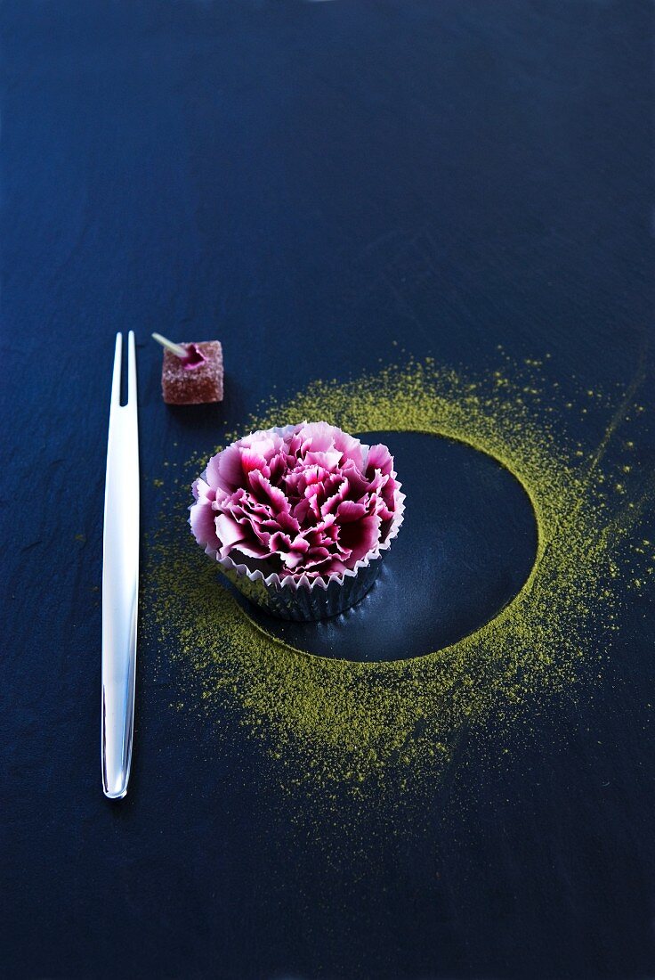 An Oriental fork next to a carnation in a muffin case and green tea powder on a black surface