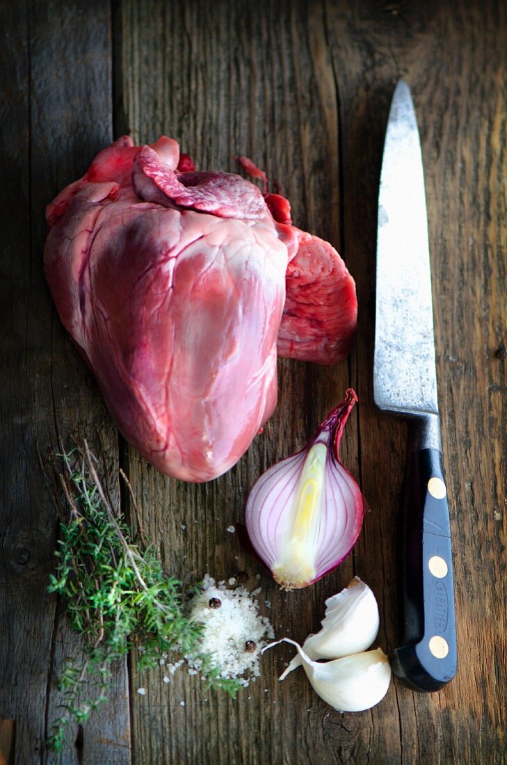 A pig's heart on a wooden board with an old knife, onions, garlic, thyme and salt