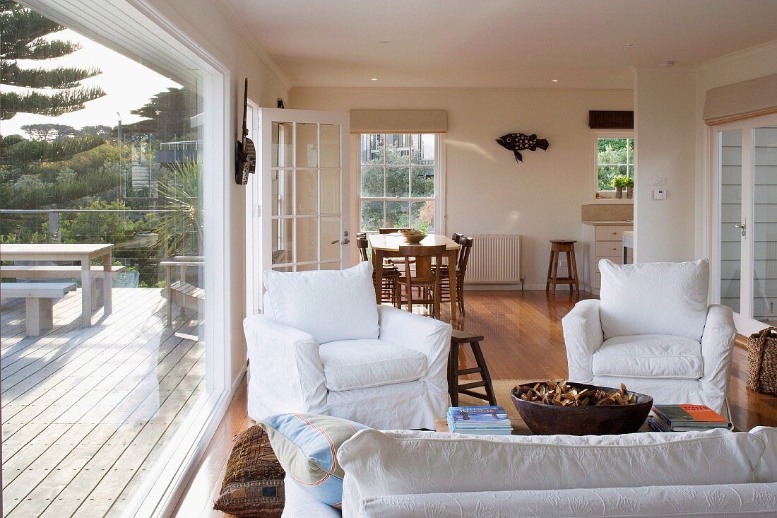 Open-plan interior of country house - armchairs with white loose covers around coffee table, dining area in background and view of furnished wooden terrace through glass wall to one side