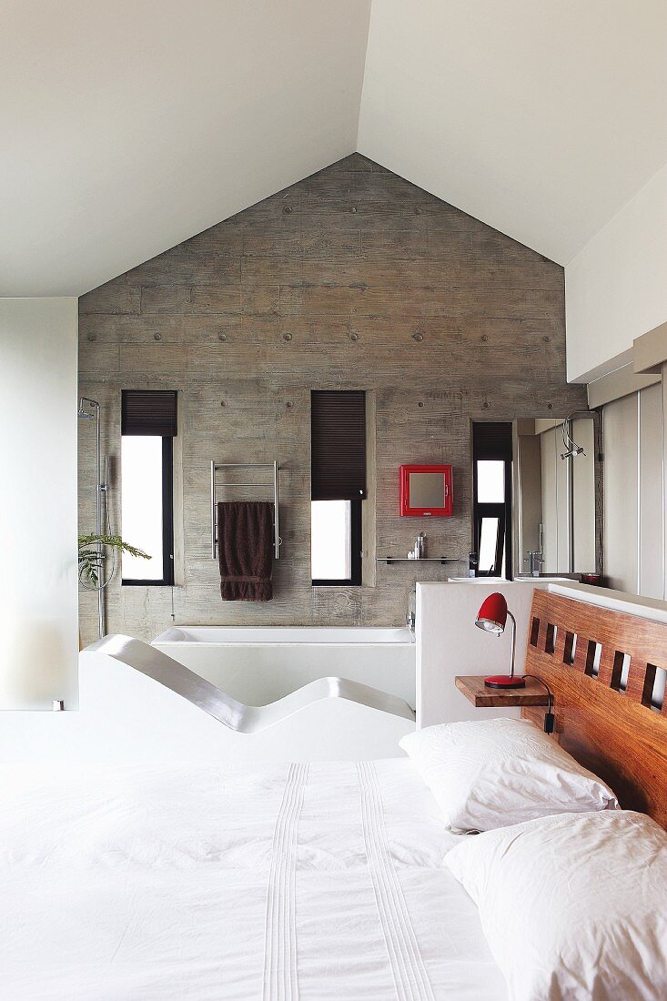 View over double bed with fitted headboard to concrete gable end wall in ensuite bathroom; integrated couch as partition