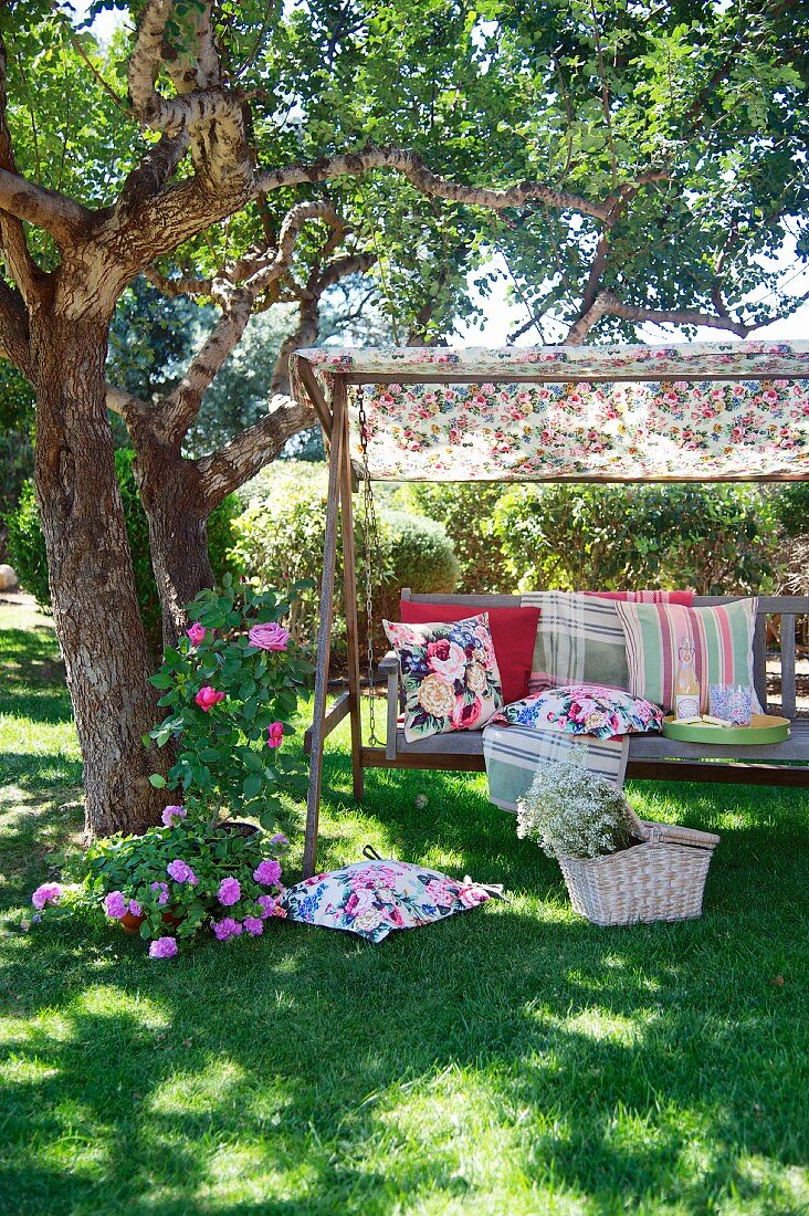 Different patterned scatter cushions on rustic garden swing seat under shady tree with picnic basket on ground