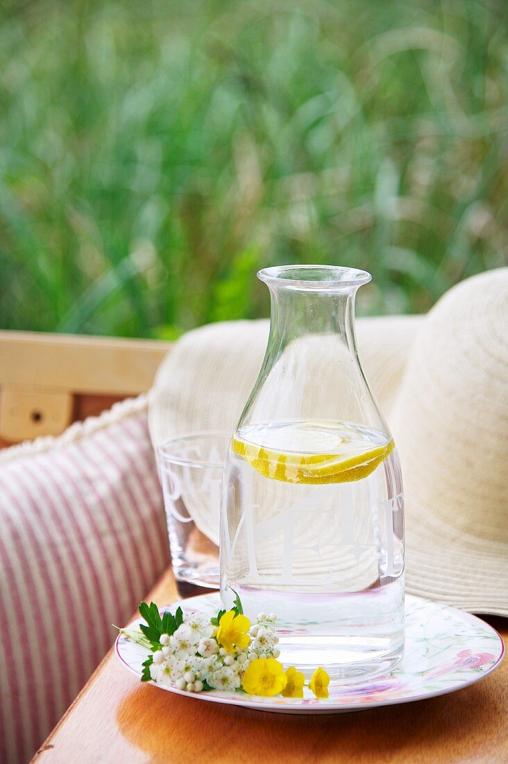 Mit Zitronenscheiben und Wasser gefüllte Flasche auf Teller mit gepflückten Wiesenblumen, dahinter teilweise sichtbarer Strohhut auf Holzunterlage