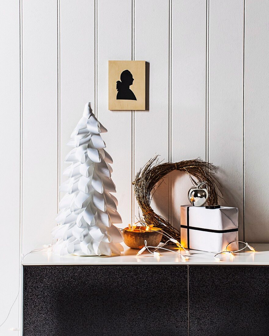 Stylised Christmas tree made from polystyrene cone covered in white ribbon strips, fairy lights and willow wreath on sideboard