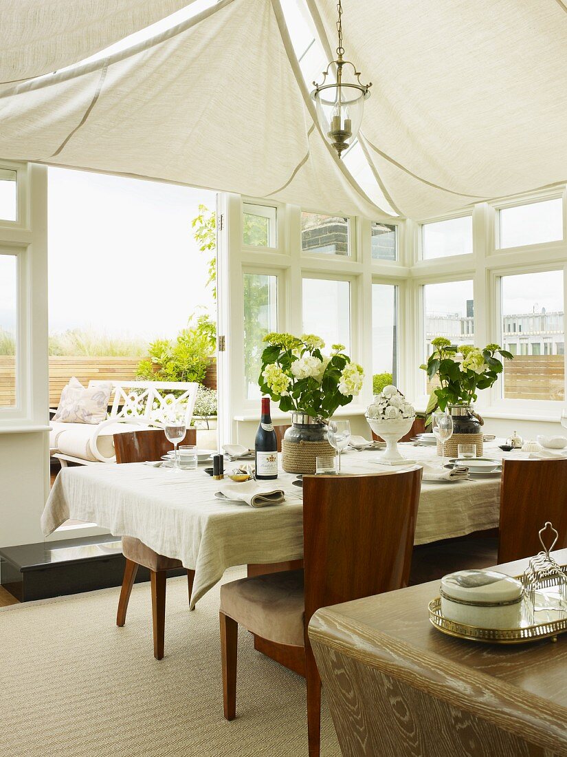 Gedeckter Tisch unter weissen Sonnensegeln an Decke im eleganten Wintergarten mit geöffneter Terrassentür