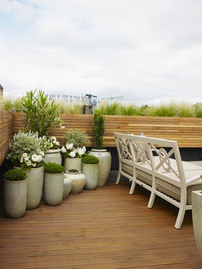 Helle bepflanzte Keramiktöpfe in der Ecke einer Dachterrasse neben weisser, traditioneller Sitzbank auf Holzdielen