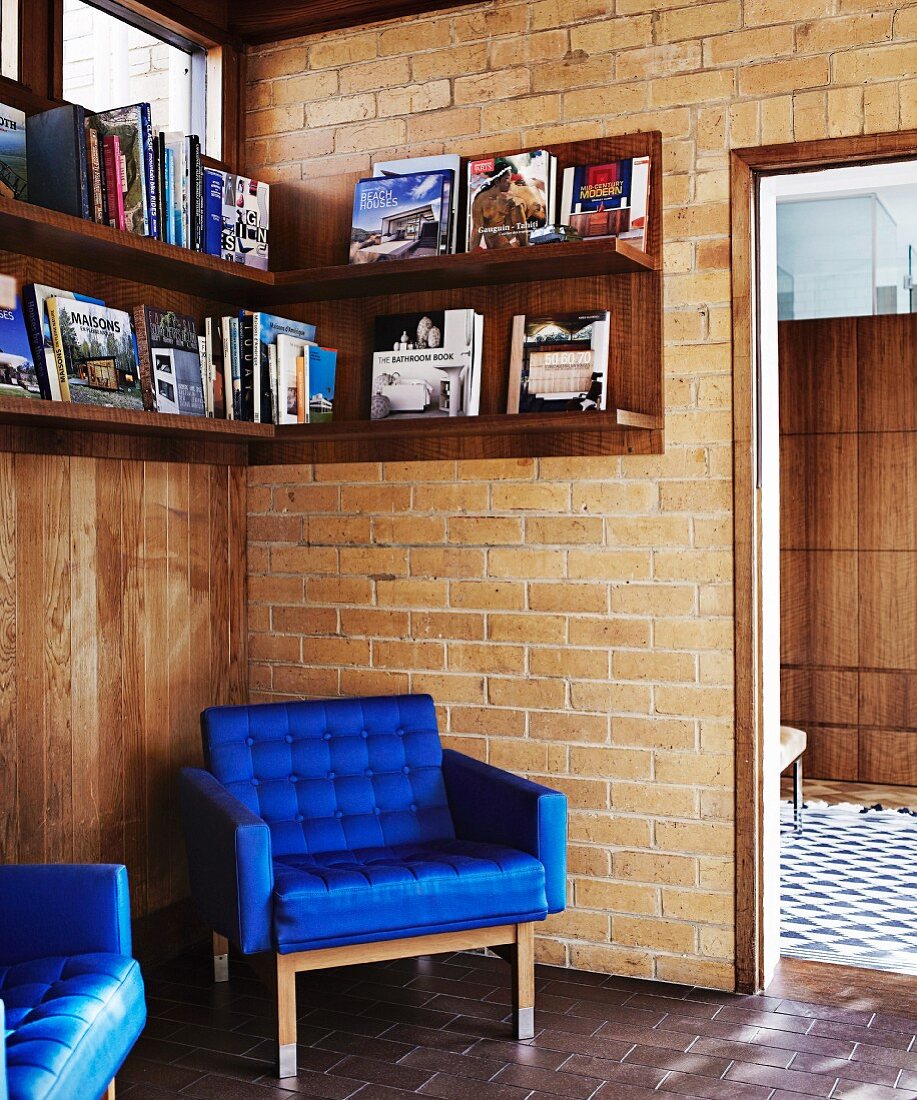 Armchair with royal blue cover in corner below wall-mounted shelves on brick wall