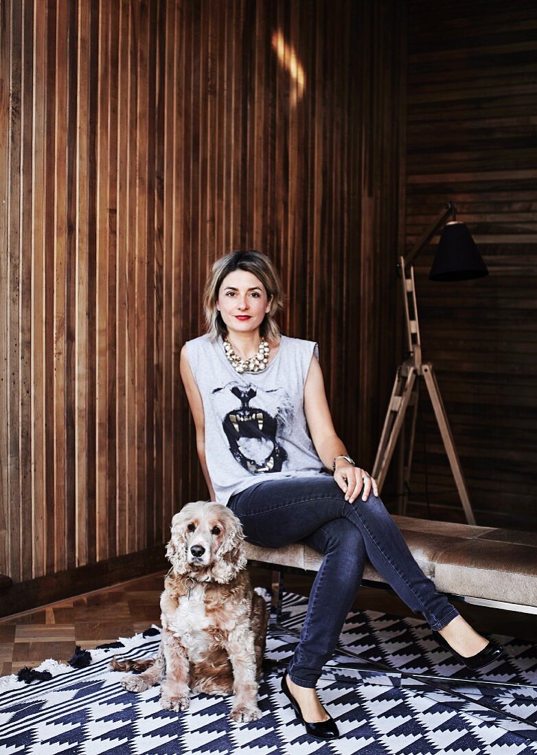 Woman sitting on bench and dog on rug with geometric pattern in front of wooden wall