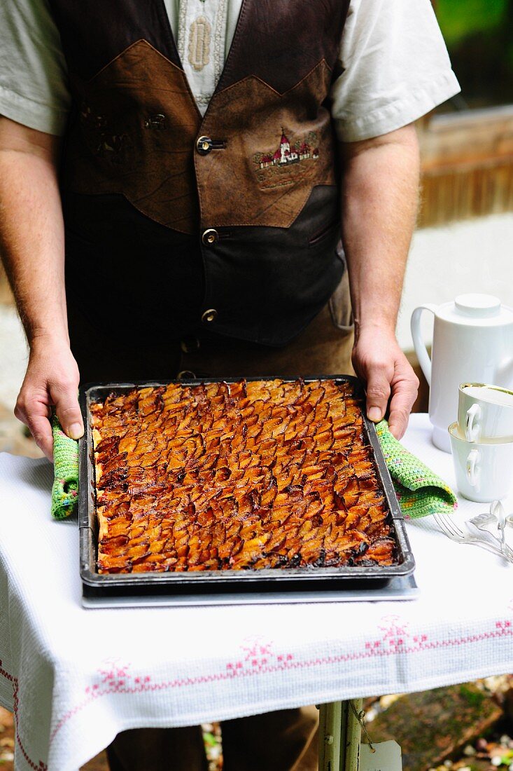 A man in a traditional Bavarian costume holding a baking tray of freshly baked Zwetschgendatschi (Bavarian plum cake)