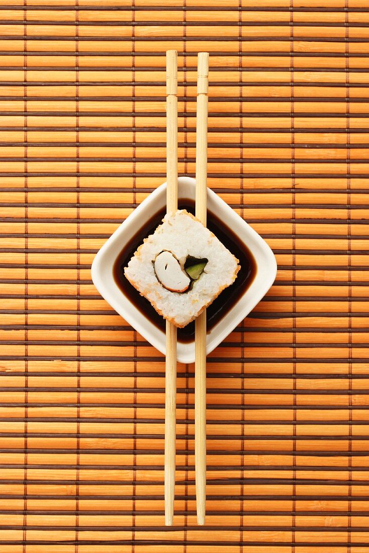 Maki sushi balanced on chopsticks over a dish of soy sauce