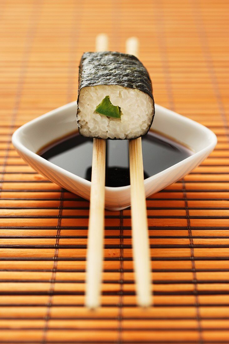 Maki sushi with cucumber balanced on chopsticks over a dish of soy sauce