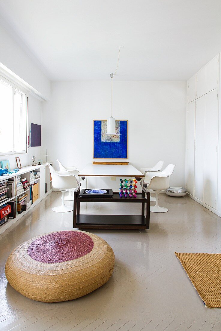 Round, basketwork floor cushion in front of black tea trolley and dining area with white classic chairs in minimalist interior