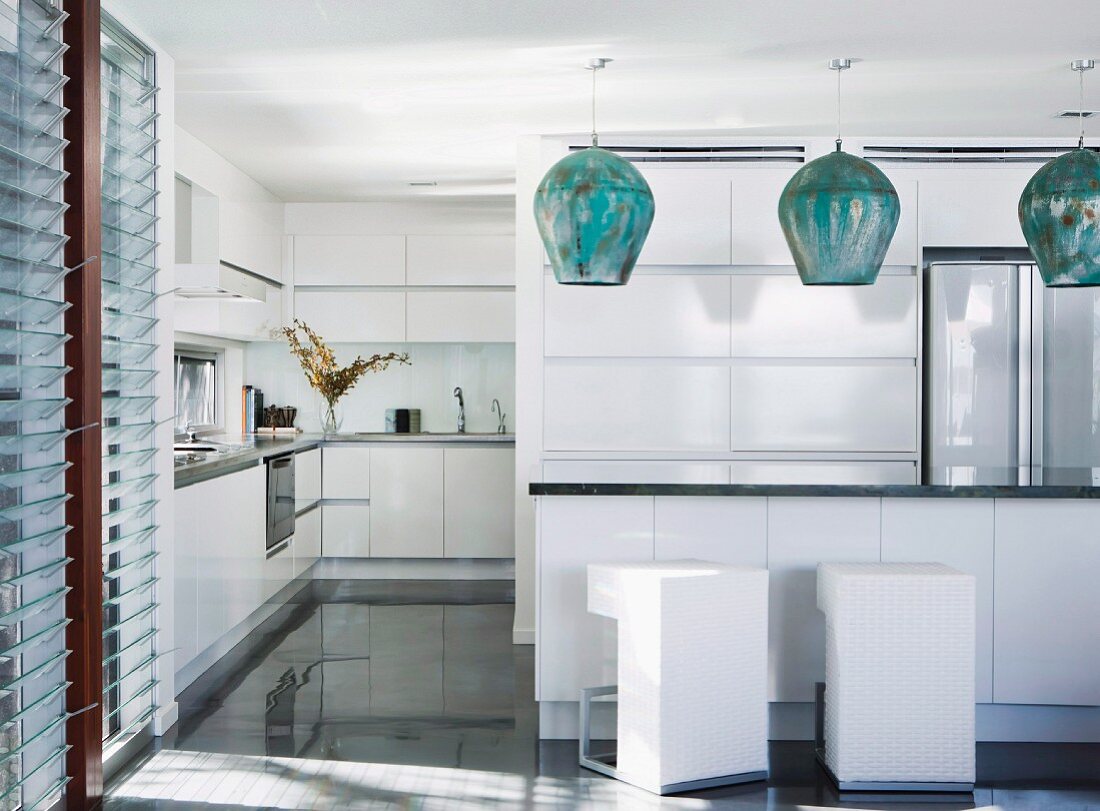 Modern white open-plan kitchen; bulbous metal pendant lamps above counter with white designer cubic bar stools