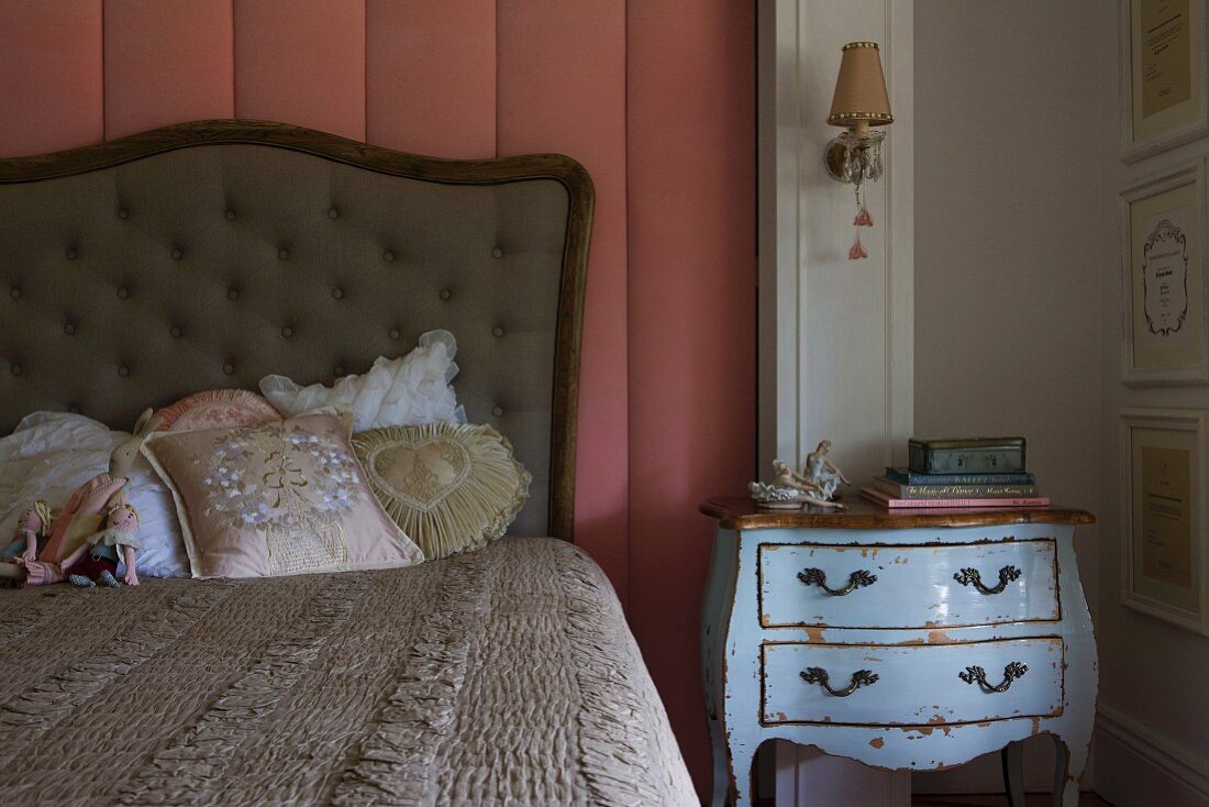 Romantic scatter cushions on bed with button-tufted headboard against wall panel with dusky-pink cover next to pale-blue battered chest of drawers