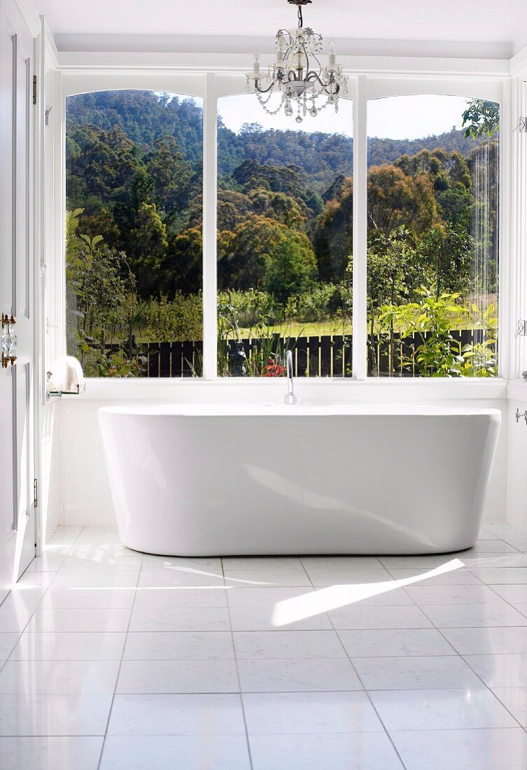 Chandelier and free-standing designer bathtub in sunny bathroom with view over woodland landscape