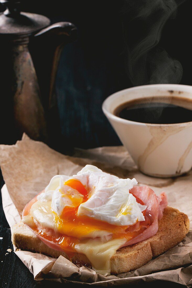 Ham and cheese on toast topped with a poached egg on a piece of baking paper served with a cup of coffee