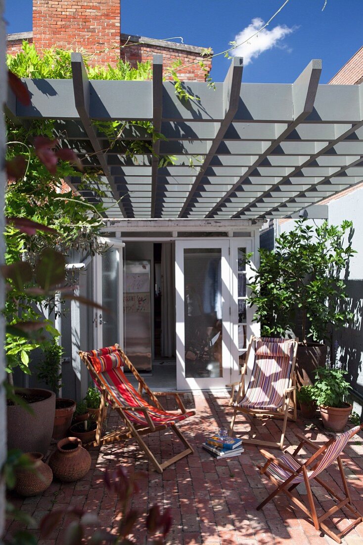 Sonnenbeschienene Liegestühle auf Terrasse, unter Pergola aus grauer Kassetten-Holzkonstruktion, im Hintergrund Wohnhaus mit offener Tür