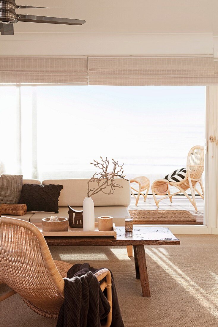 Helles, freundliches Wohnzimmer mit Rattansessel, hellem Sofa und rustikalem Couchtisch vor Panoramafenster