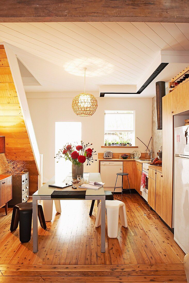 Vase of flowers on modern table in open-plan kitchen, rustic wooden floor, white-painted wooden ceiling and staircase in background