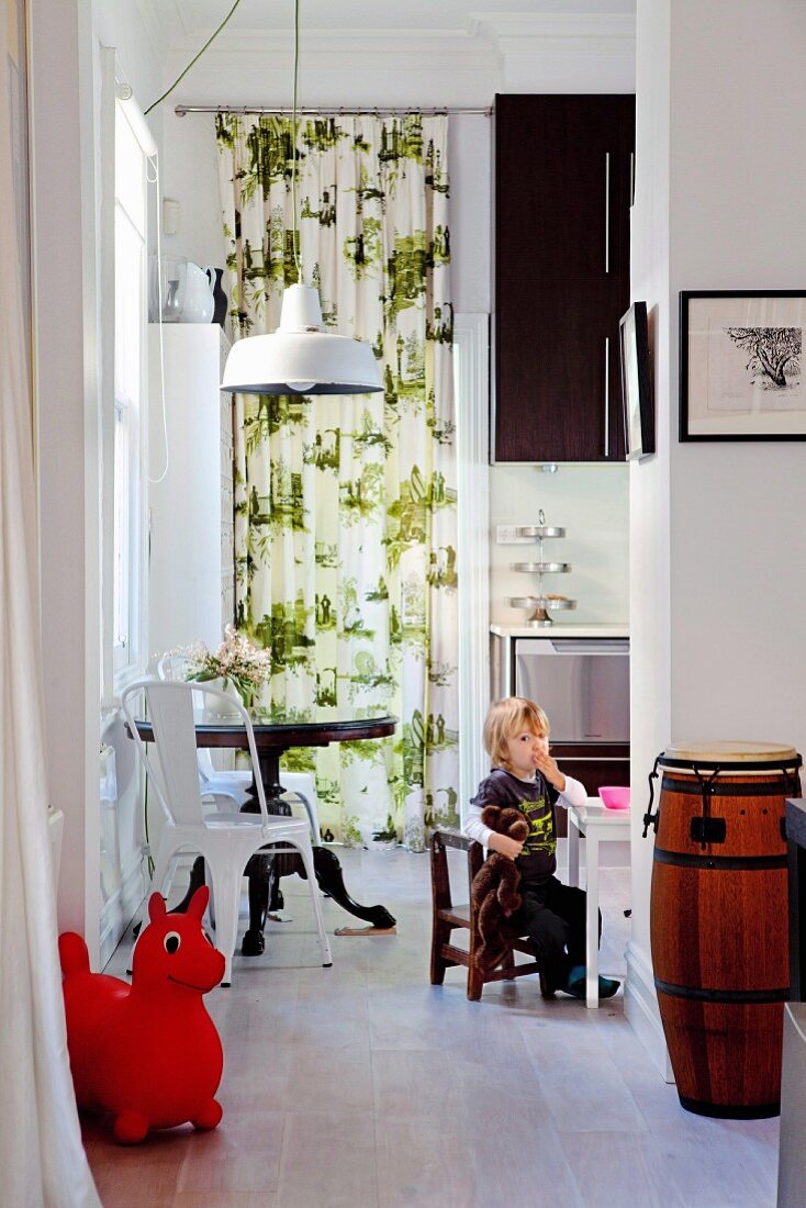 Young child sitting on child's chair in kitchen, retro chair at round table and floor-to-ceiling patterned curtain