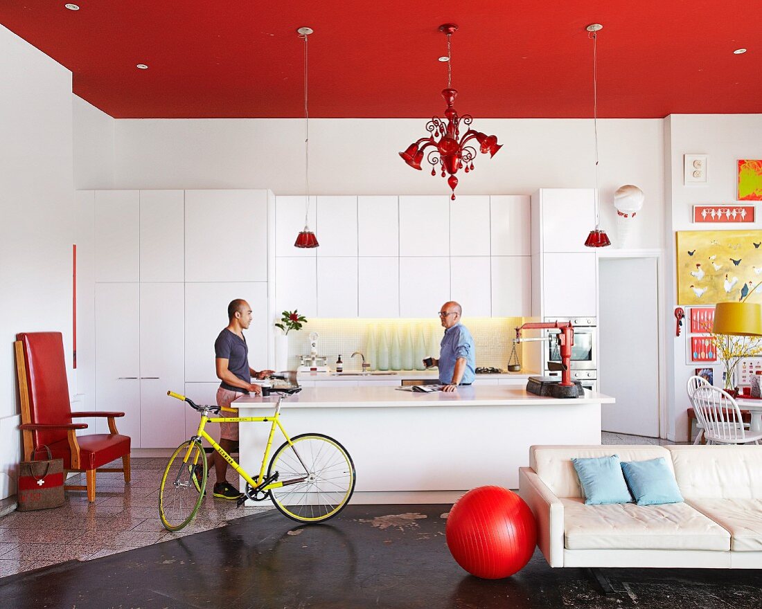 Open-plan interior with red ceiling above white kitchen area and men next to free-standing island counter; red ball next to white, designer sofa in foreground