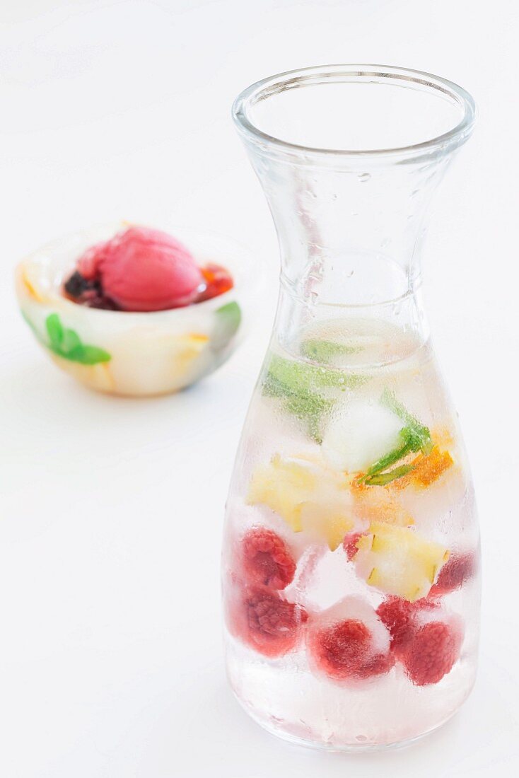A carafe of water and fruit ice cubes with a bowl of raspberry ice cream in the background