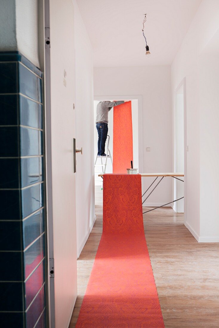 Red, patterned wallpaper rolled out on floor and table; view of man hanging wallpaper in background