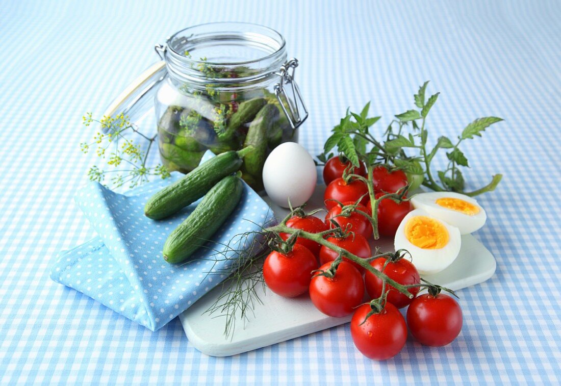 A board and a napkin with tomatoes, eggs, cucumbers and herbs and a jar of gherkins on a blue and white checked tablecloth