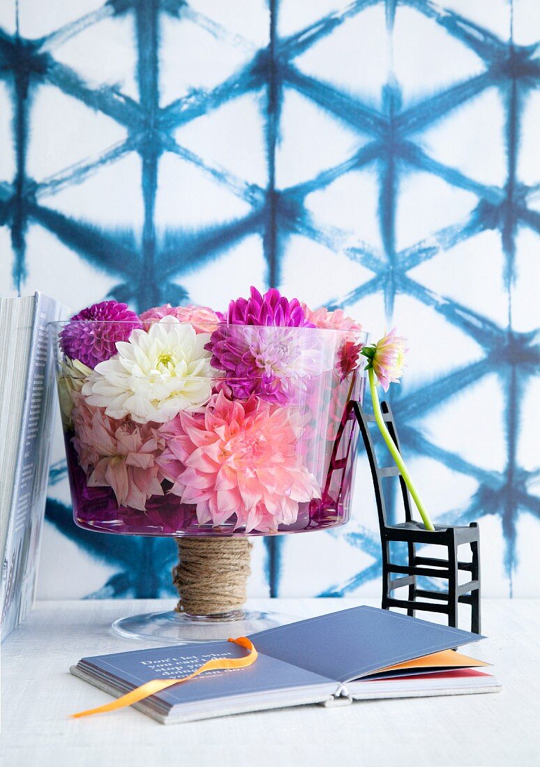 Glass bowl of dahlia flowers, black chair ornament and book in front of wall with blue mesh pattern