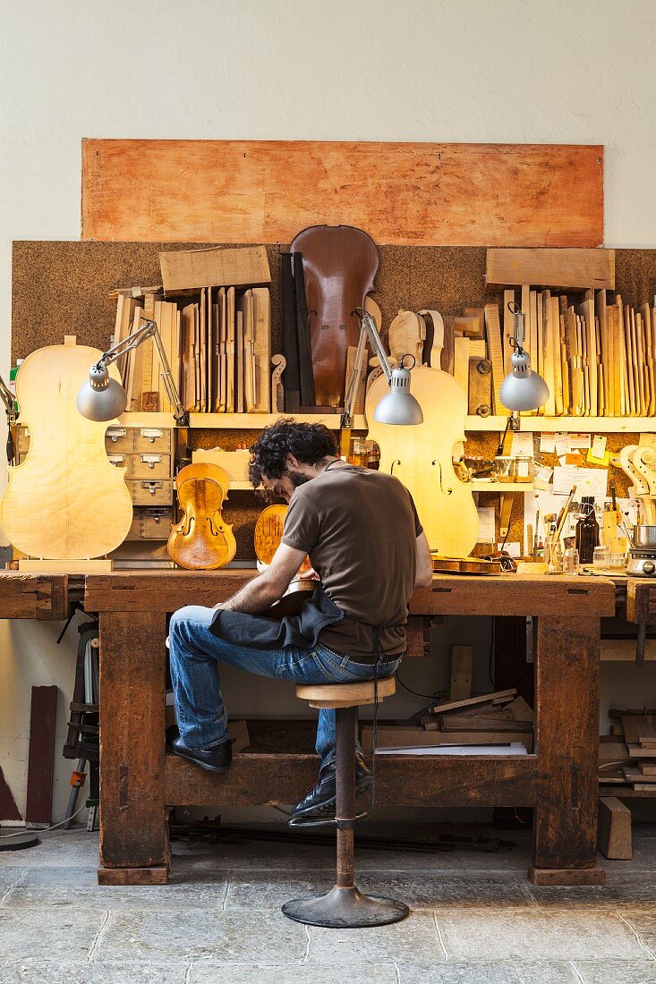 Violin-maker sitting in front of half-finished instruments on illuminated workbench