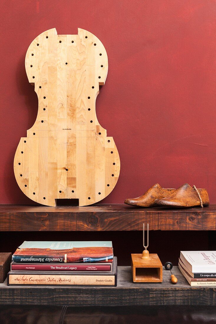 Rustikale Holzablage mit Bücherstapel und Stimmgabel, Schuh-Modelle aus Holz und heller Cellokorpus an dunkelrote Wand gelehnt