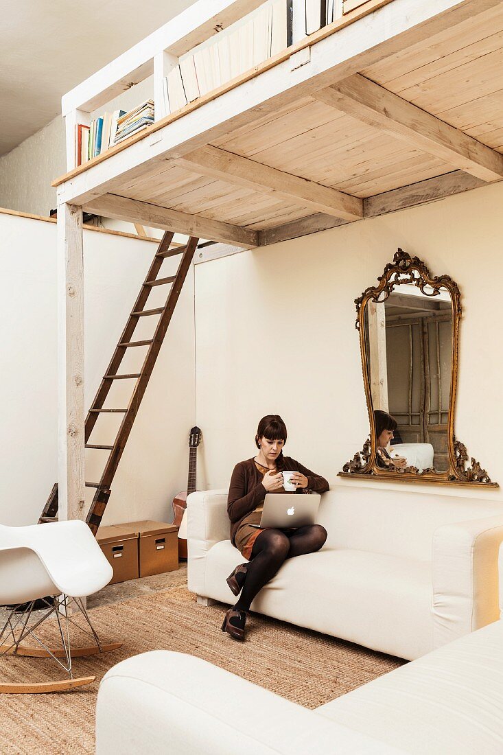 Young woman with laptop sitting on ecru sofa below white-painted wooden mezzanine accessed by wooden ladder in open-plan interior with vintage character