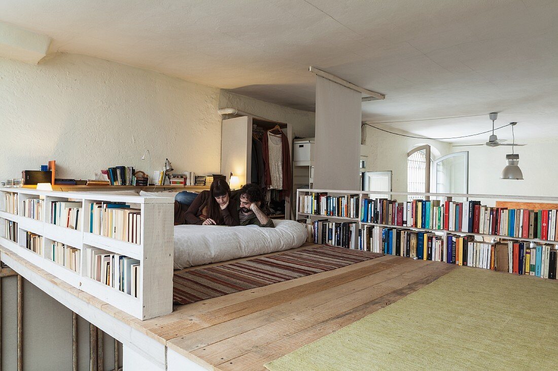 Young couple reading on wooden mezzanine surrounded by books on balustrade bookshelves