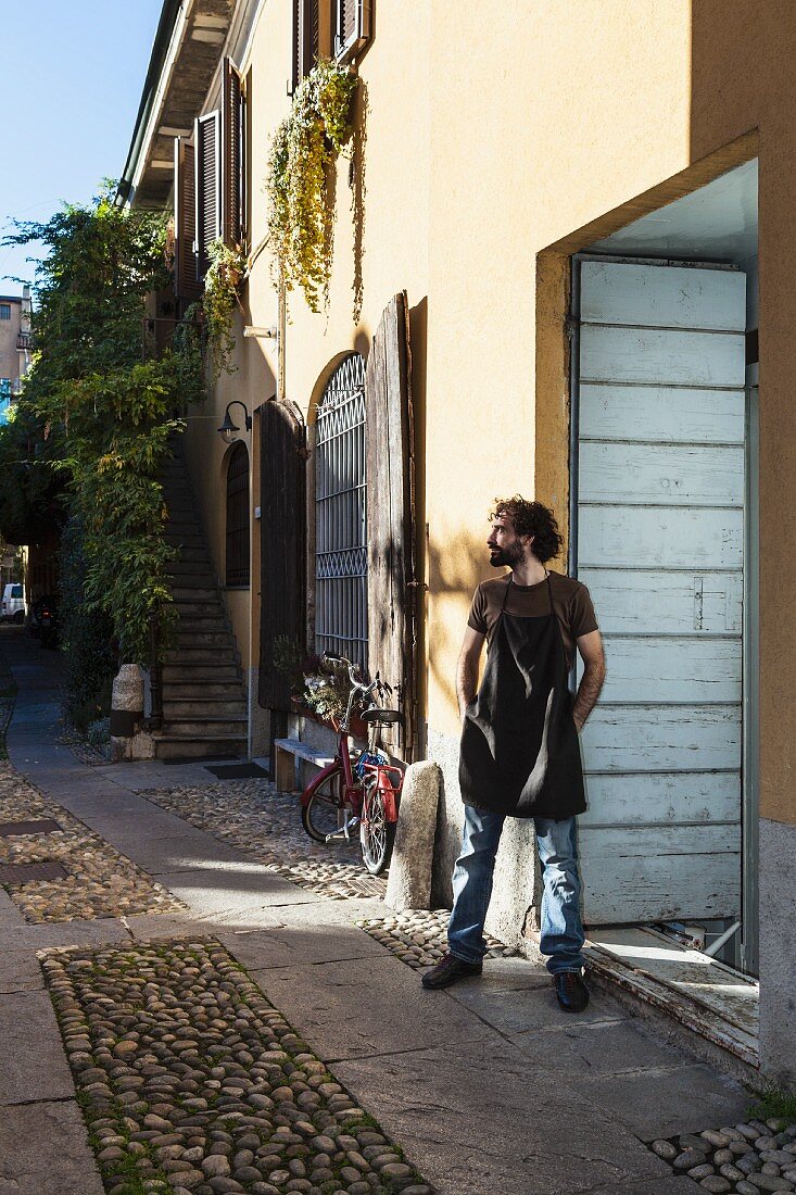 Italienischer Geigenbauer mit Arbeitsschürze an Hauswand vor Werkstatt gelehnt