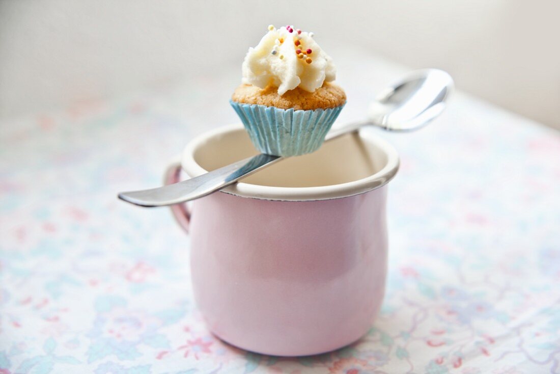 A mini cupcake balanced on a spoon over a pink enamel mug