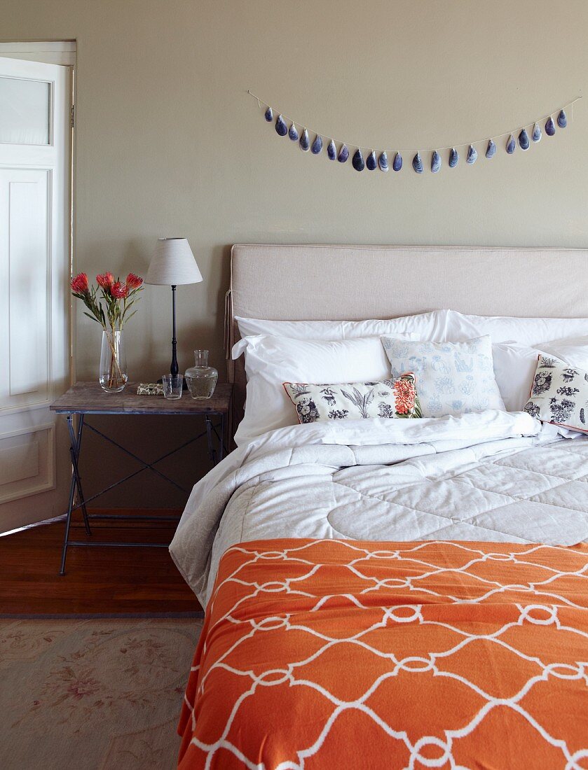 Double bed with headboard, orange bedspread with ogee pattern and rustic bedside table in simple bedroom