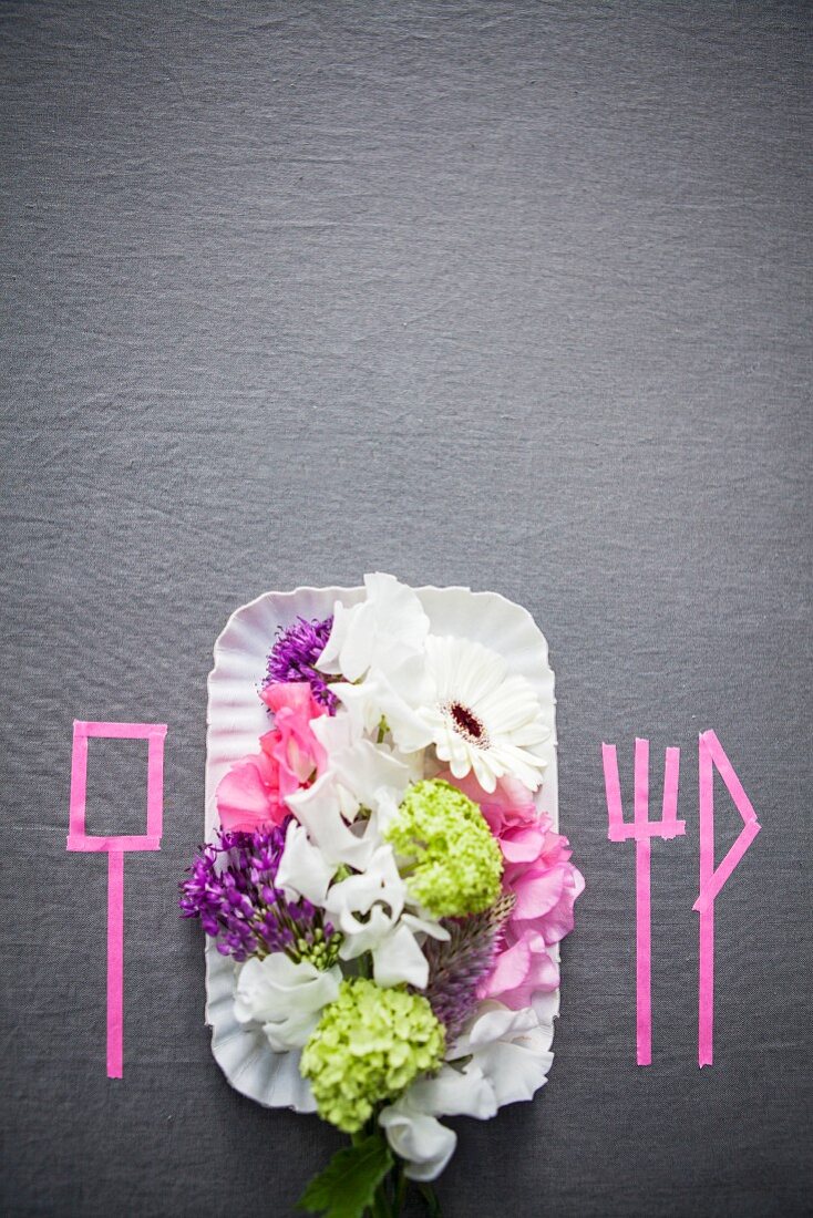 Place setting with summer flowers and silhouettes of cutlery marked on grey surface with pink washi tape