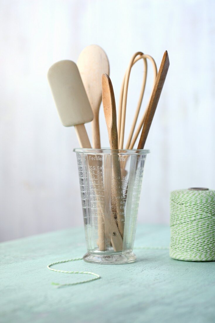 Various wooden kitchen utensils in a measuring jug next to a roll of green kitchen twine