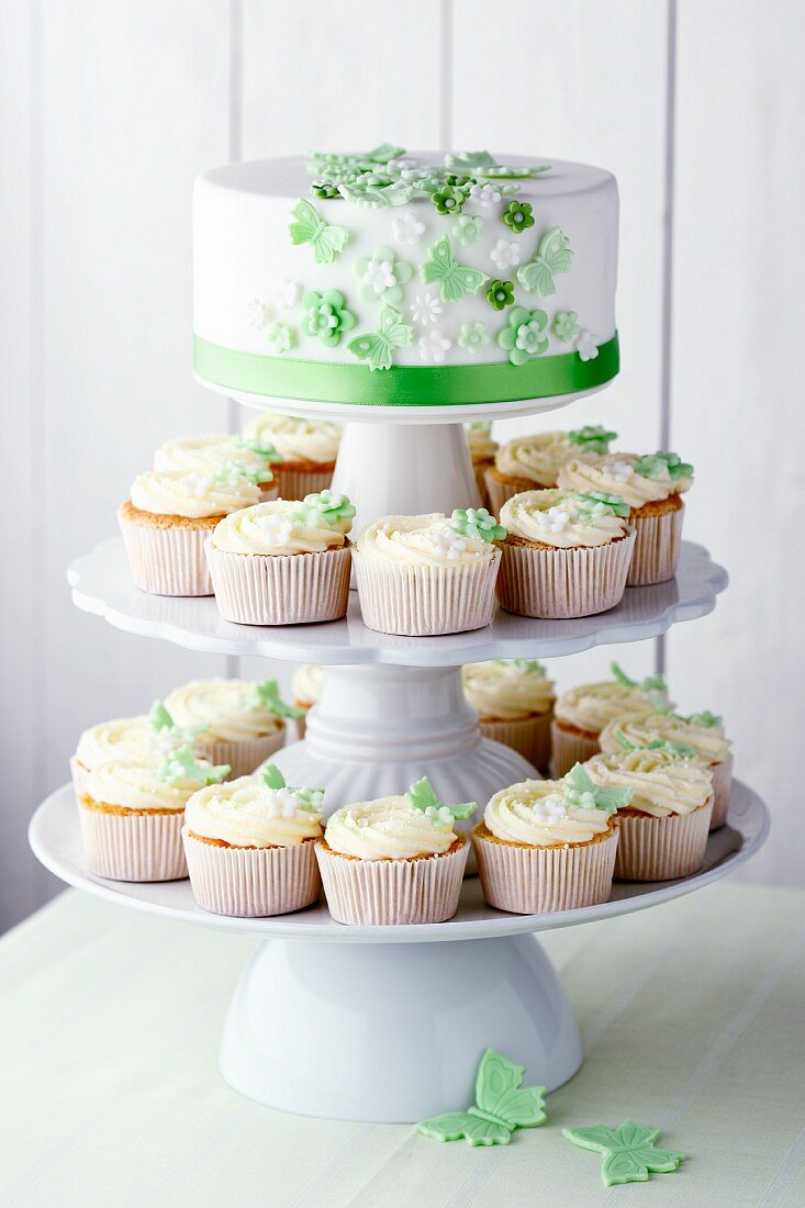 Mehrere Vanille-Cupcakes und Hochzeitstorte mit grünen Zuckerschmetterlingen und Zuckerblüten auf Etagere