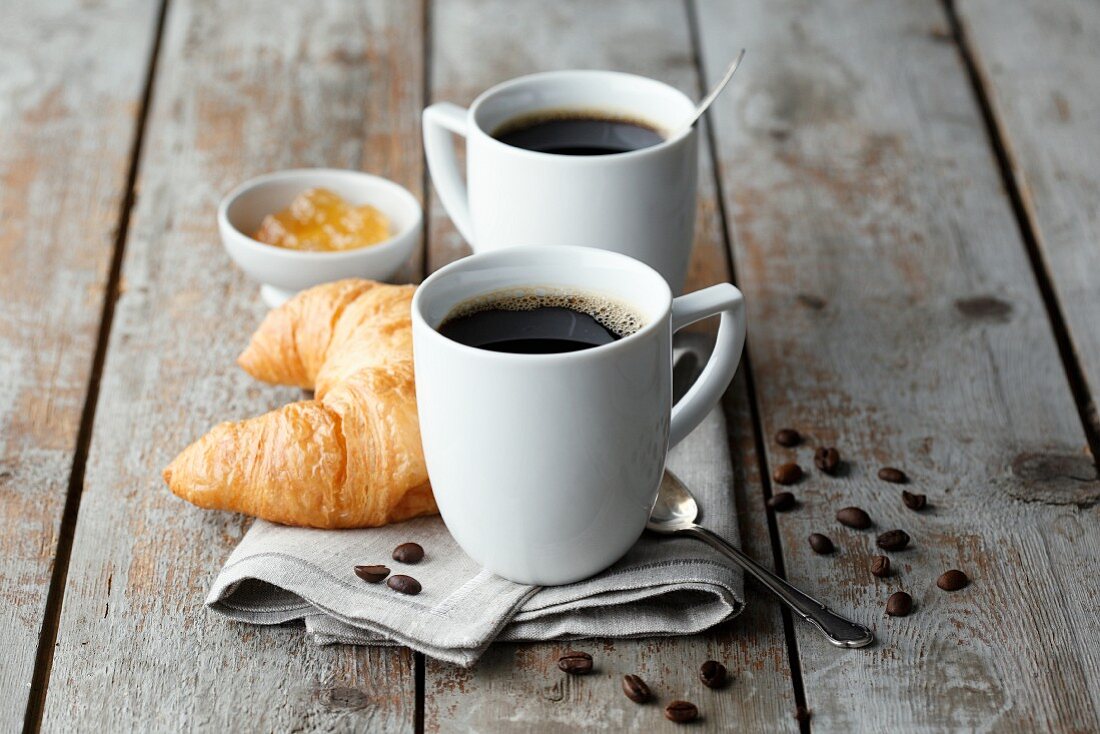 Mugs of coffee on a napkin with coffee beans served with a croissant and jam