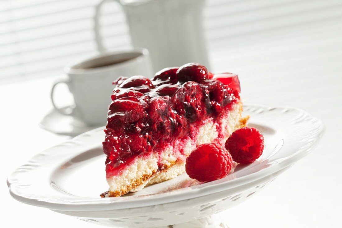 A slice of raspberry cake on a cake stand and with a coffee pot and a cup of coffee in the background