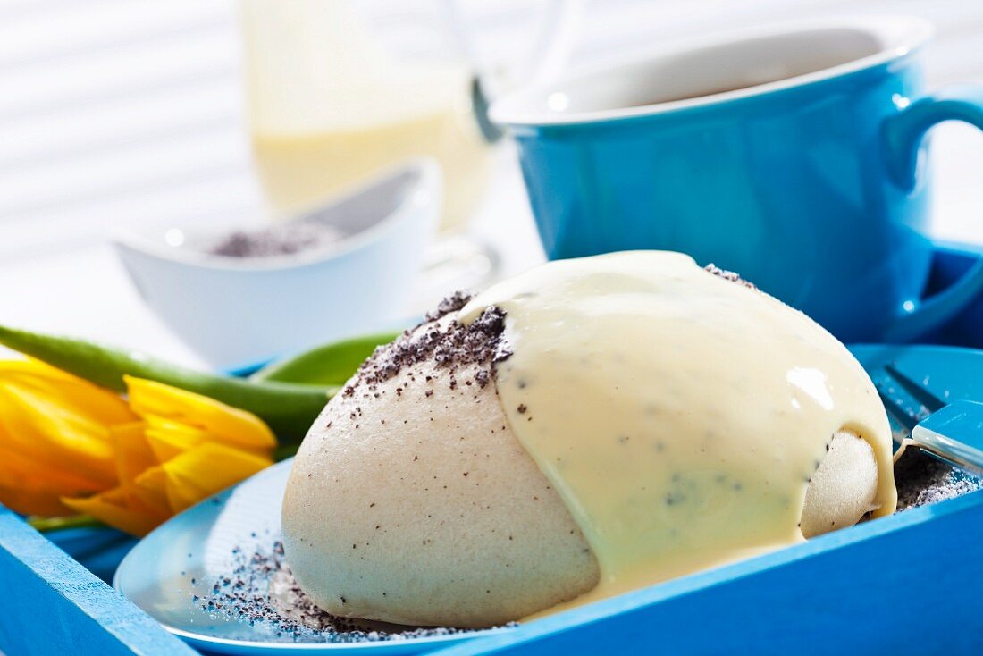 A yeast dumpling topped with poppyseeds and vanilla sauce served with a cup of coffee and a tulip