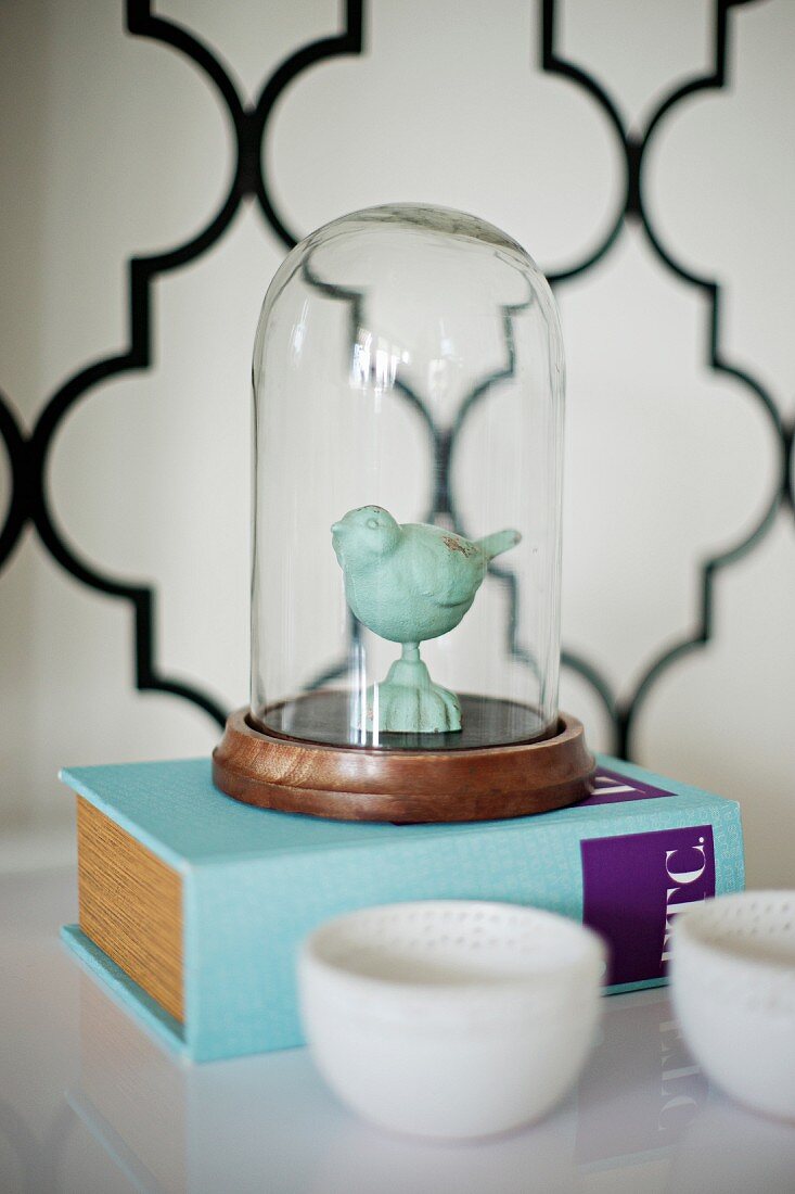 Bird figurine under glass cover on thick book; wallpaper with Oriental tile pattern in blurred background