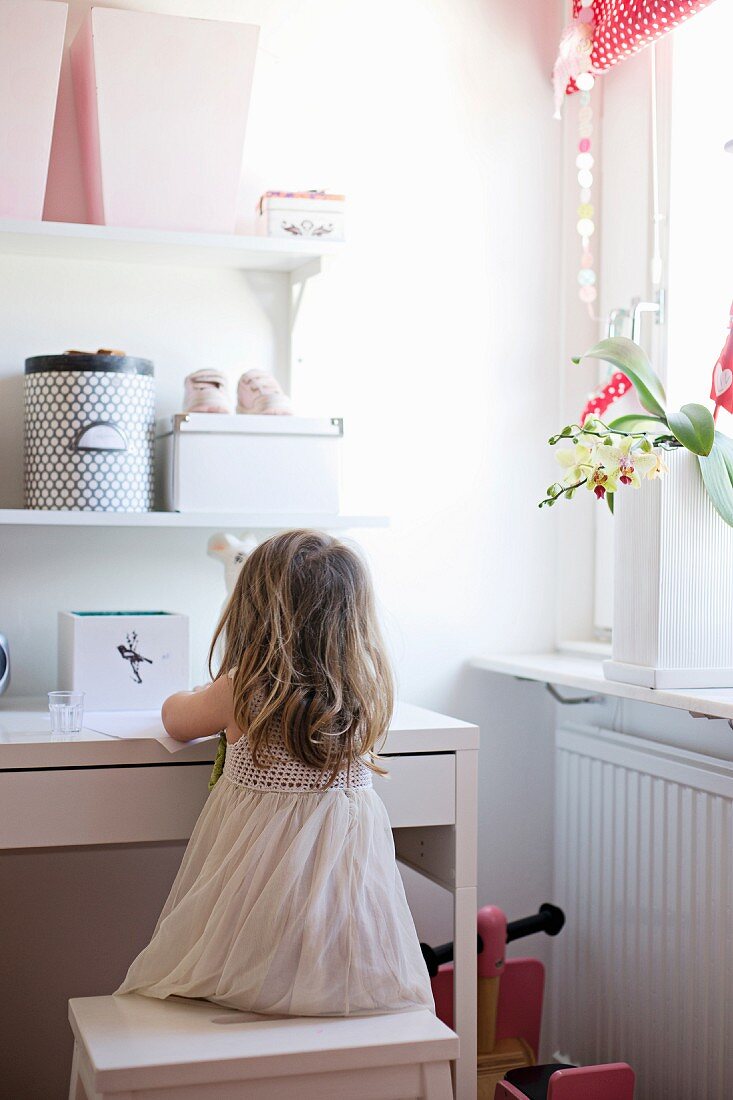 Kleines Mädchen auf Hocker vor weißem Schreibtisch und Wandborden mit Aufbewahrungsboxen
