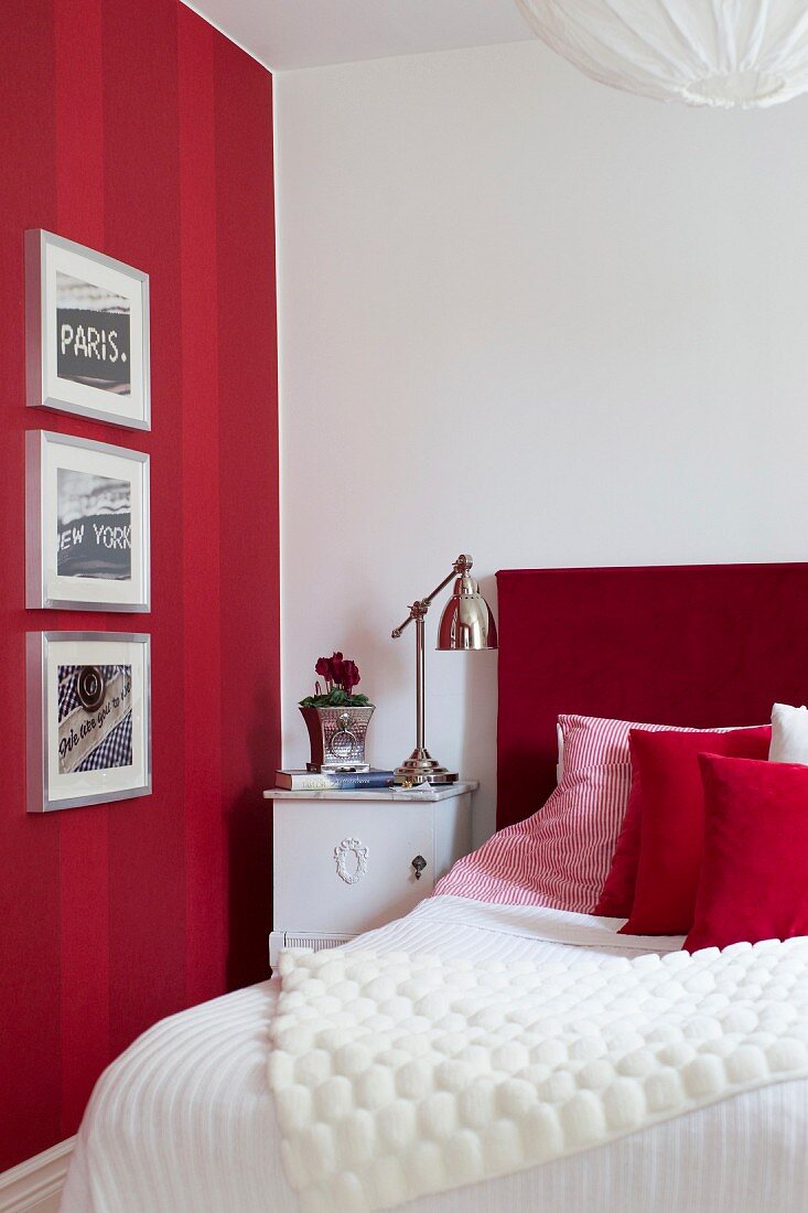 Gerahmte Bilder an rot gestreifter Wand, daneben ein Bett mit rotem Kopfteil und Leuchte im Retrostil auf Nachtkasten