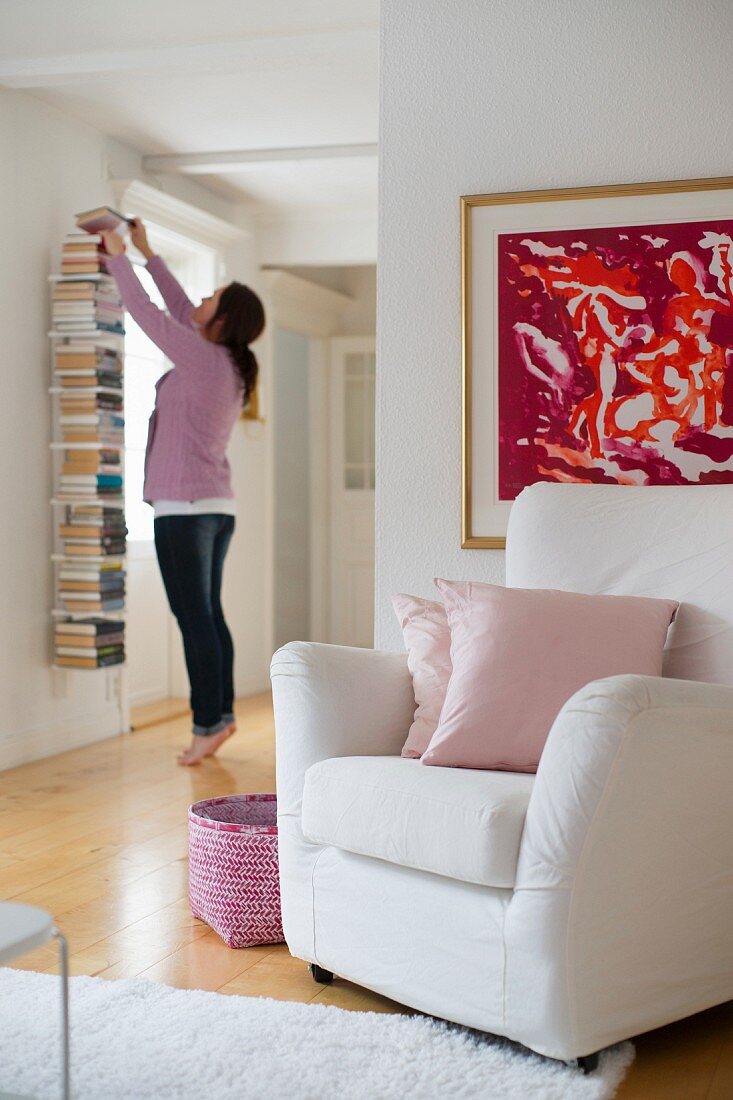 Modern artwork above white armchair and woman in front of bookshelf in open-plan hallway