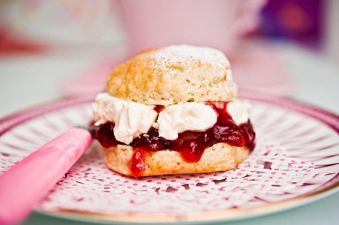 A scone with cream and strawberry jam on a doily with a teacup in the background