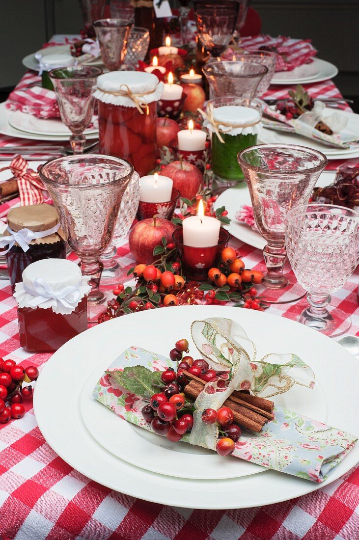 A Christmas table laid with a red checked tablecloth, candles, napkins and jars of jam